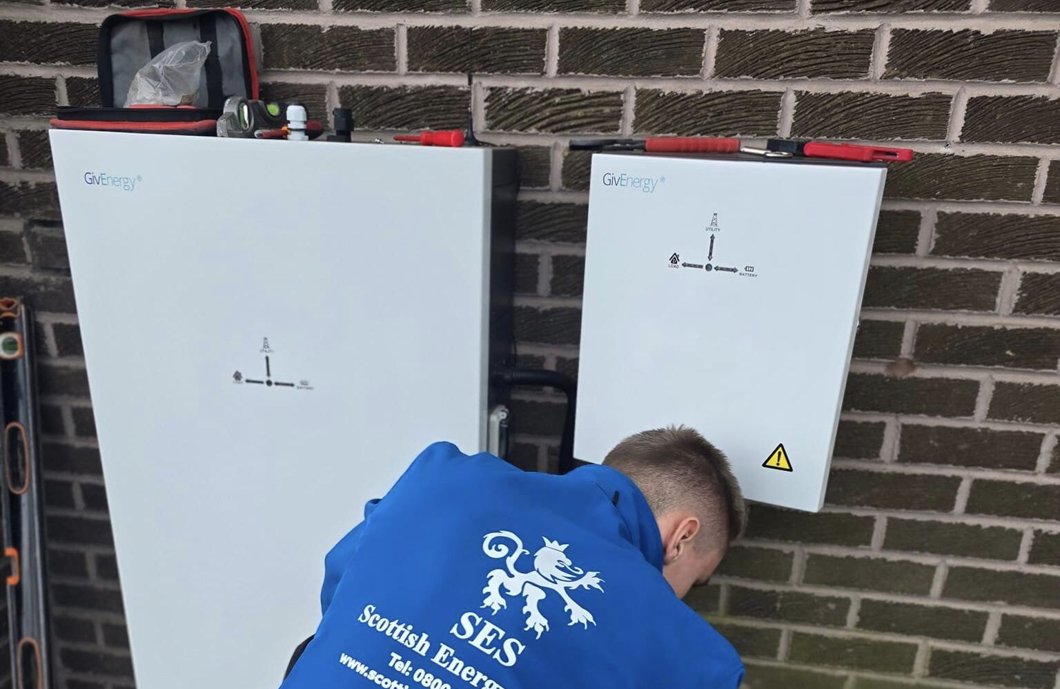 An engineer in a blue SES Scottish Energy jacket working on the installation of GivEnergy battery systems mounted on a brick wall, with tools and equipment visible nearby.