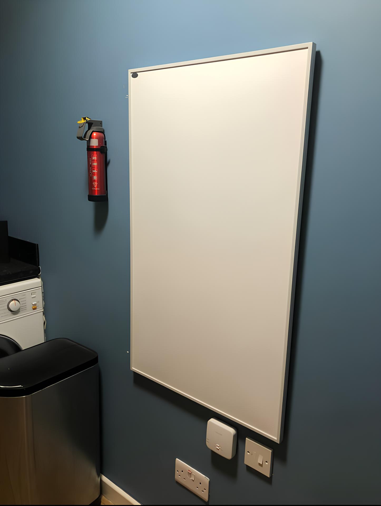Photo of a large white electric panel heater mounted on a wall in a utility room. A fire extinguisher is mounted on the wall next to the heater.