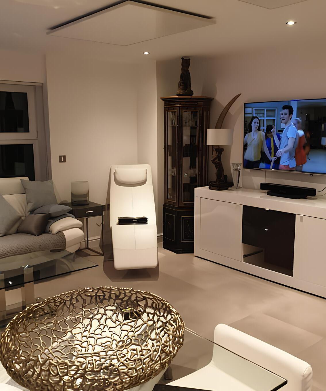 Photo of a white infrared heater installed on the ceiling of a modern living room. The heater is mounted above a glass coffee table and a white sofa.