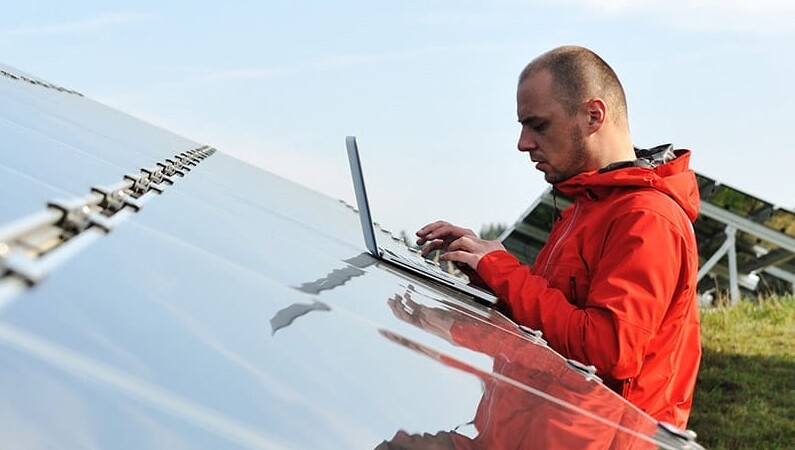 A technician in a bright red jacket inspecting solar panels outdoors using a laptop, with a clear sky and green landscape in the background.